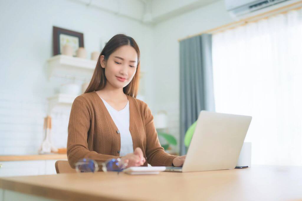 Teacher working on a laptop at a desk in a bright home office while using a calculator, with a calm and focused expression. This image represents Singapore Math Teacher Training and Singapore Math professional development of Mindset 4 Math, highlighting an educator preparing lessons or building instructional expertise using the CPA approach and bar model method.